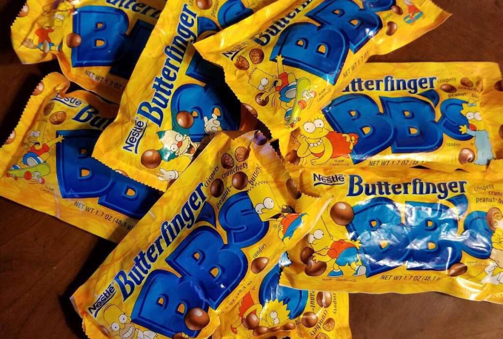 A pile of yellow bags of Butterfinger BB's candy is spread out on a wooden surface. The packaging features cartoon characters and blue text with images of small, round chocolate candies.