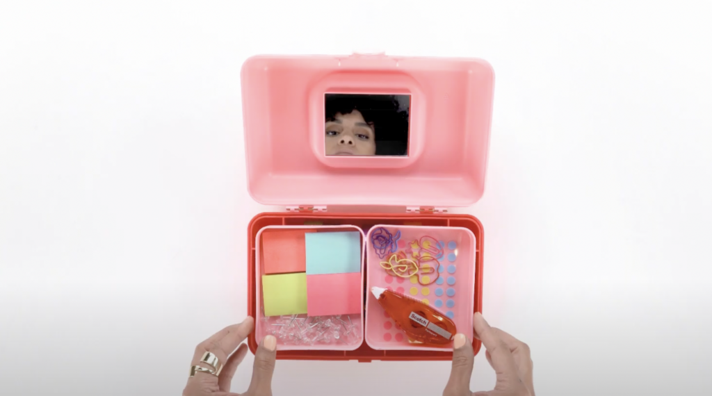 A person peers into a opened pink box, which is held by their hands. Inside the box are colorful sticky notes, paper clips, a bottle of glue, and other small office supplies, all neatly organized.