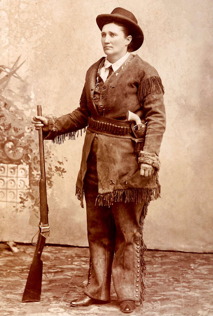 A person dressed in 19th-century frontier attire, including a fringed jacket, holds a rifle. They wear a wide-brimmed hat and have a belt with a holster and ammunition. The sepia-toned image has a floral backdrop.