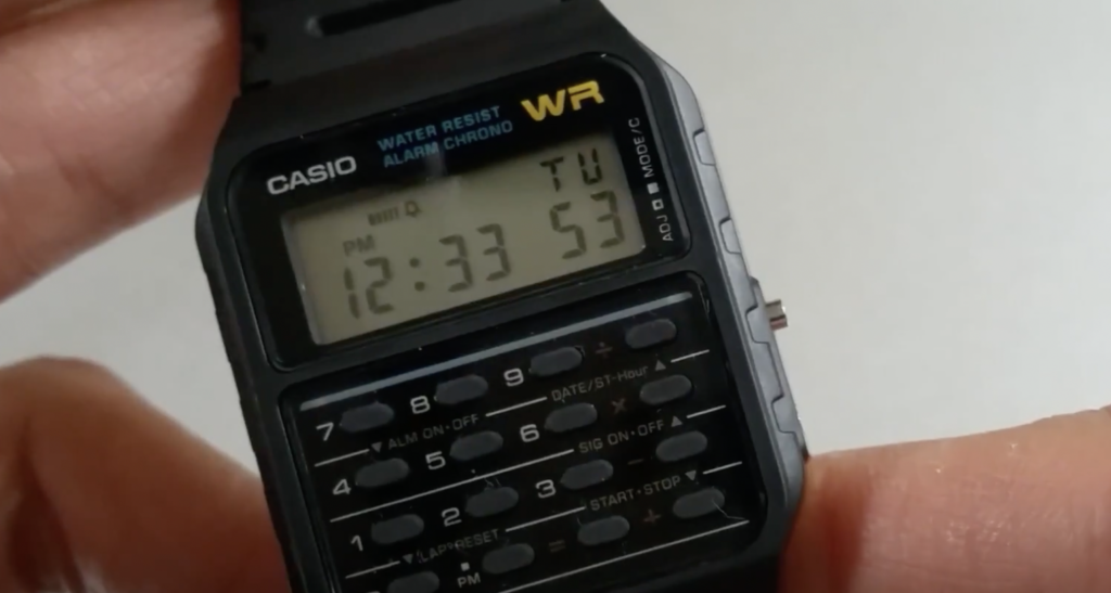 Close-up of a black Casio digital watch with a calculator keypad. The display shows the time as 12:33 and the date as Tuesday the 5th. The watch features multiple buttons for various functions.