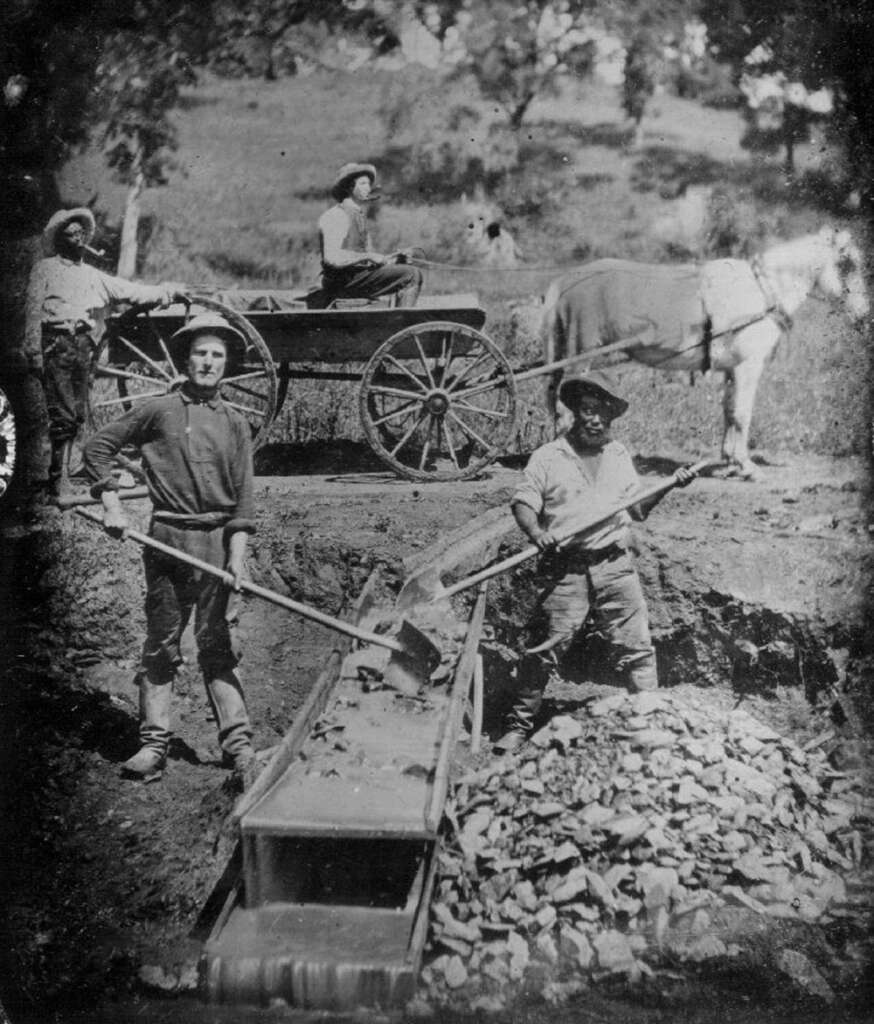 Historical black-and-white image showing four men during the 19th-century Gold Rush. Two men dig at a sluice box filled with rocks, while another stands by a horse-drawn cart in the background. Trees and grassy terrain are visible.