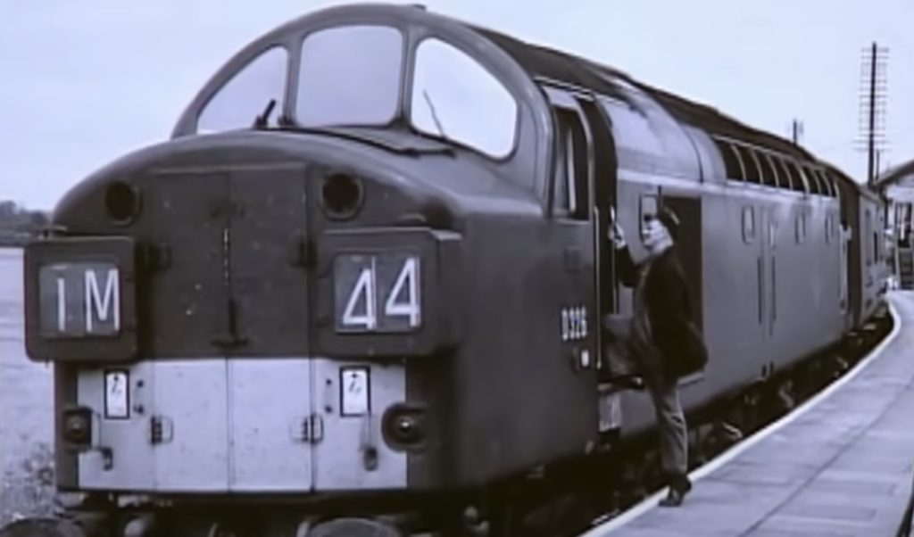 A vintage train with the number 44 on the front is stopped at a station platform. A person is standing on the steps, partially entering the train. The scene is in black and white.