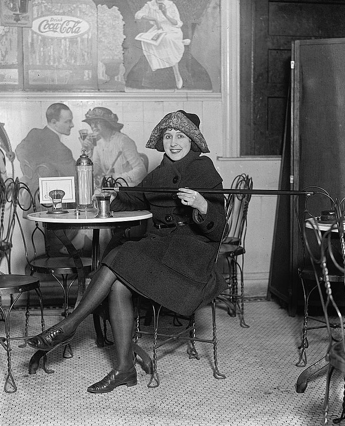 Vintage photo of a woman sitting at a small café table, wearing a coat and hat. She holds a spoon and appears to be stirring her drink. Behind her is a large painting of a Coca-Cola advertisement with two people.