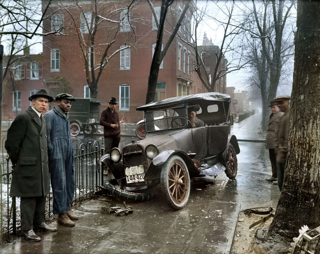 A vintage car with a damaged wheel is parked on the side of a wet, tree-lined street. Five men stand around, wearing hats and coats, observing the scene. Buildings in the background suggest an urban neighborhood.