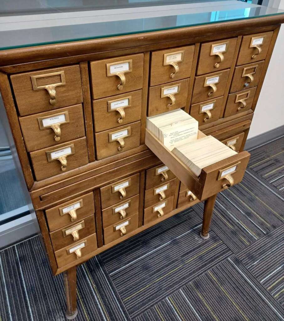 A wooden card catalog with multiple drawers, commonly used in libraries, stands against a glass wall. One drawer is open, revealing index cards inside. The floor is carpeted with a striped pattern.