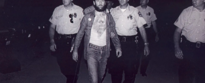 A man with a beard wearing a T-shirt and jeans is being escorted by four uniformed police officers. They are walking forward in a dimly lit setting. The police officers are wearing hats and badges.