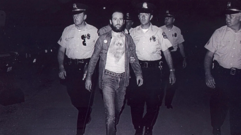 A man with a beard wearing a T-shirt and jeans is being escorted by four uniformed police officers. They are walking forward in a dimly lit setting. The police officers are wearing hats and badges.