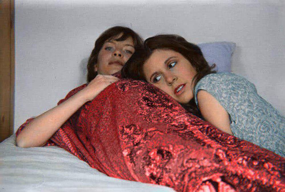 Two women are lying on a bed. One is under a red blanket, while the other, wearing a light blue top, leans her head against the first woman's shoulder. Both appear relaxed and contemplative.