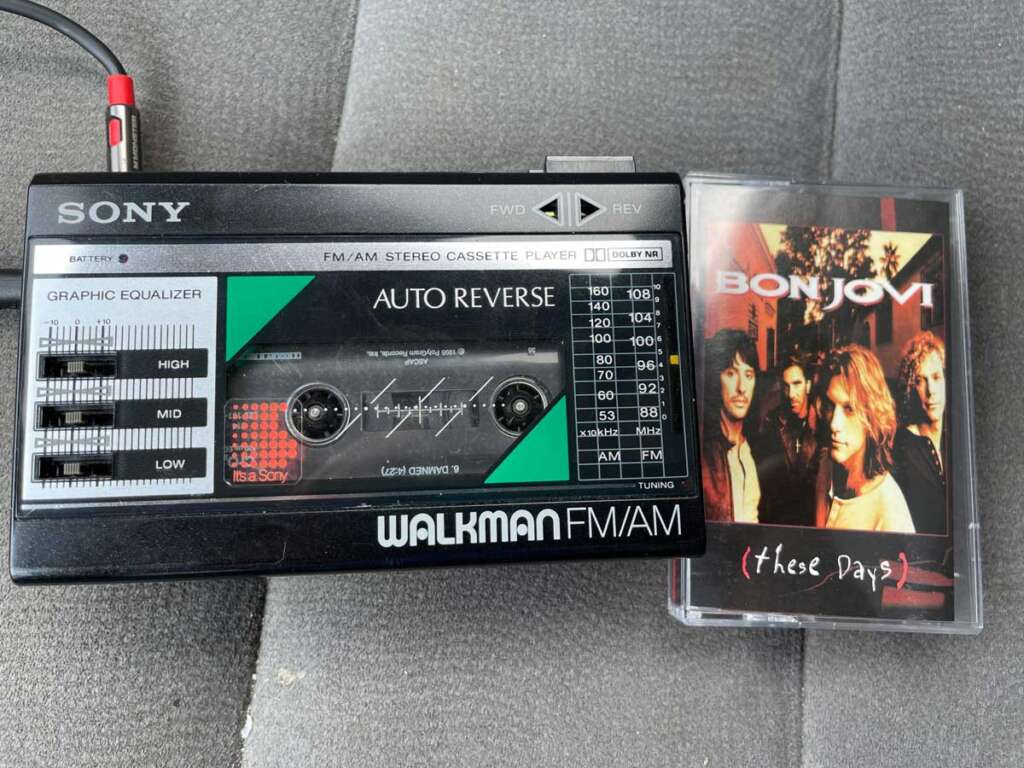 A vintage Sony Walkman with a radio tuner and cassette tape inside, placed next to a Bon Jovi "These Days" cassette tape cover featuring band members on its front. The Walkman has a graphic equalizer with sliders for high, medium, and low frequencies.