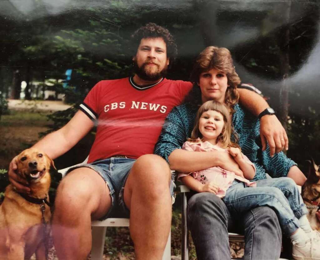 A family sits outdoors on chairs, surrounded by trees. A man in a red T-shirt holds a dog, sitting beside a woman in a patterned shirt. A young girl sits on the woman's lap, smiling. Another dog is partially visible on the right.