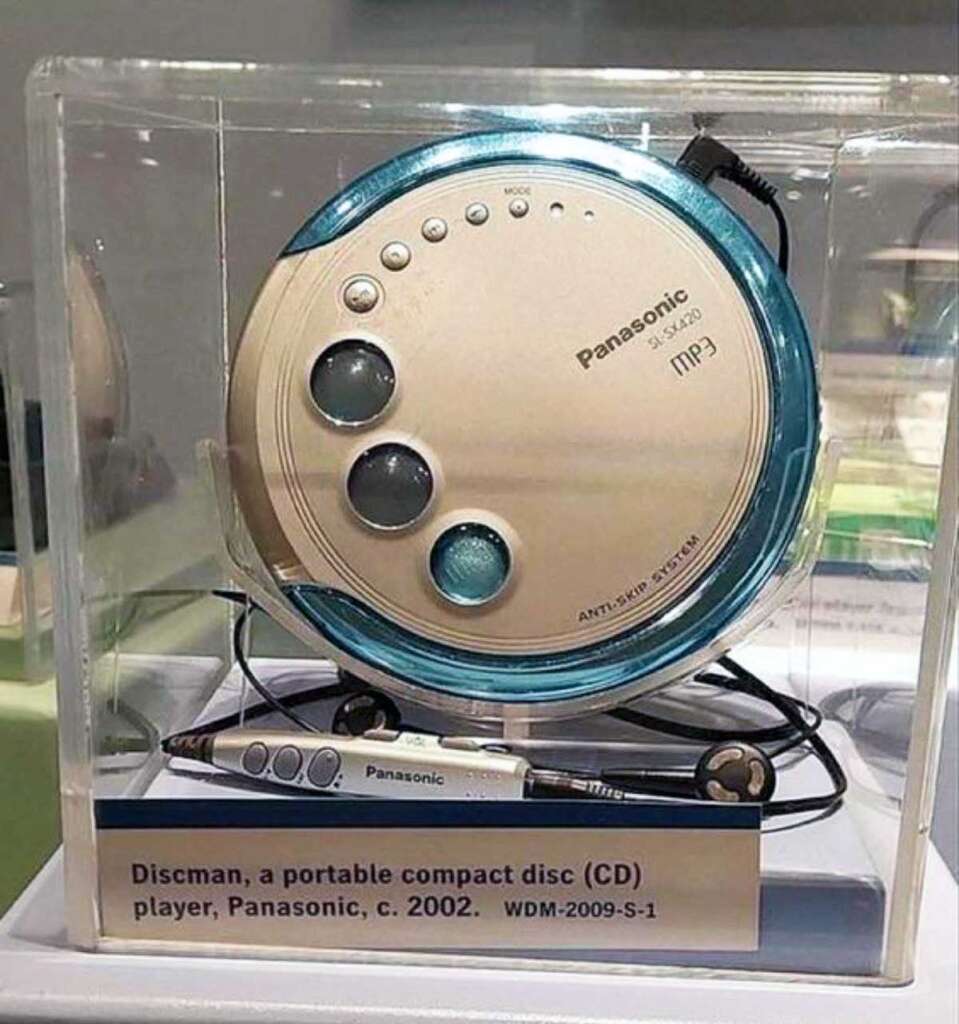 A Panasonic portable CD player with a silver and blue circular design, displayed in a clear case with attached black and silver headphones. The label reads, "Discman, a portable compact disc (CD) player, Panasonic, c. 2002. WDM-2009-S-1.