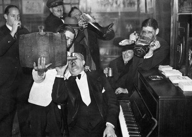 A group of men in suits and hats are gathered around a piano, drinking from bottles, mugs, and a barrel. One man plays the piano while others enthusiastically pour drinks. The setting appears lively and jovial.