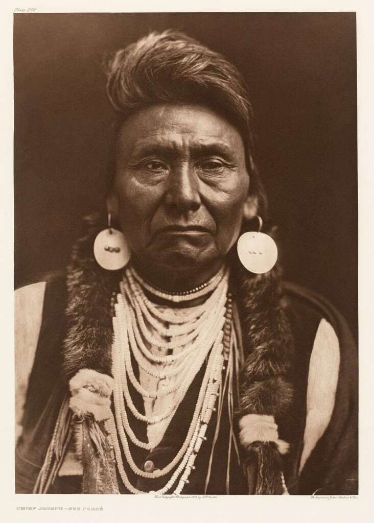 A portrait of Chief Joseph of the Nez Percé. He wears traditional attire, including large earrings and multiple beaded necklaces, with fur accents on his shoulders. His expression is serious, and the background is dark.