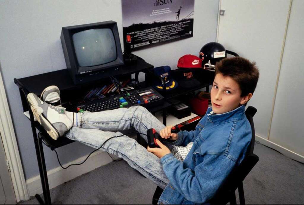 A young person sits in a chair playing video games on a retro computer setup. They hold a joystick, with their feet up on the desk. The desk has a computer monitor, keyboard, and caps, while a movie poster hangs on the wall.