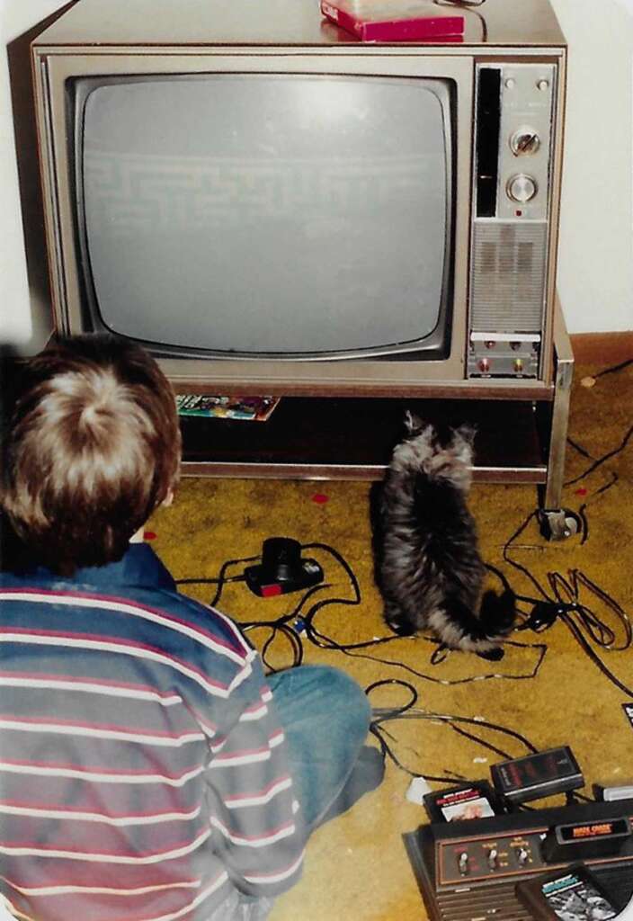 A child and a cat sit on a carpeted floor facing an old television. The screen displays a simple maze game. An Atari console with controllers and cables is scattered in front of them. The room has a cozy, retro atmosphere.