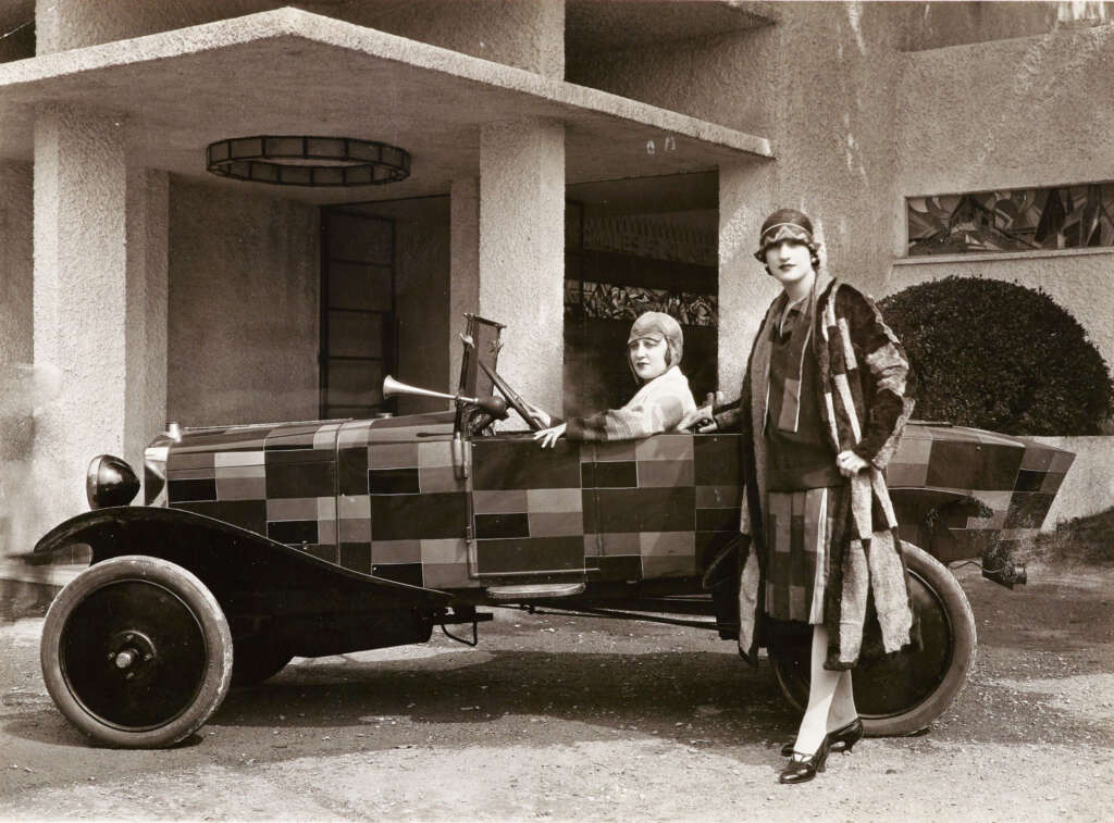 Two women dressed in 1920s attire stand beside and in a vintage car with a checkered pattern. The background features a textured building with geometric design elements.