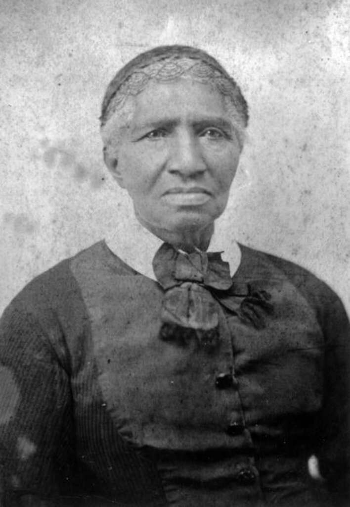 An elderly woman from the 19th century, wearing a dark dress with a high collar and a bow. Her hair is pulled back, and she appears serious, looking slightly to the side. The photo is in black and white and has an aged appearance.