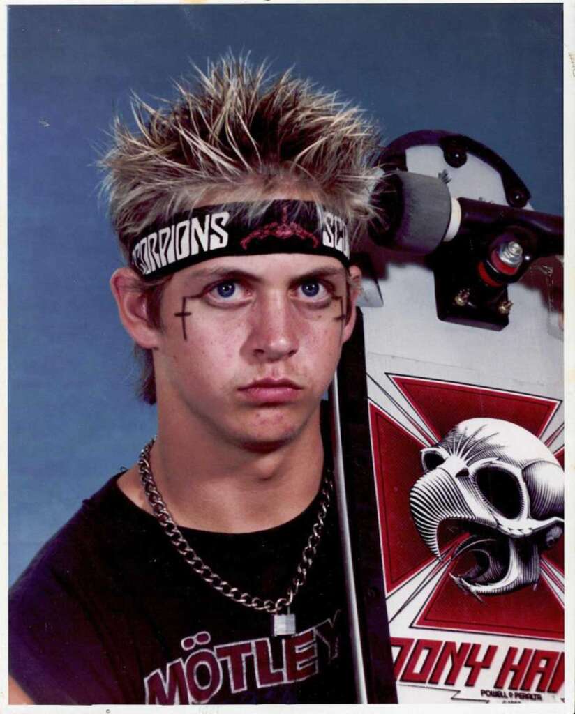 A young person with spiked hair and face paint stands holding a skateboard featuring a skull design. They wear a black band T-shirt, a bandana, and a chain necklace. The background is a plain blue color.