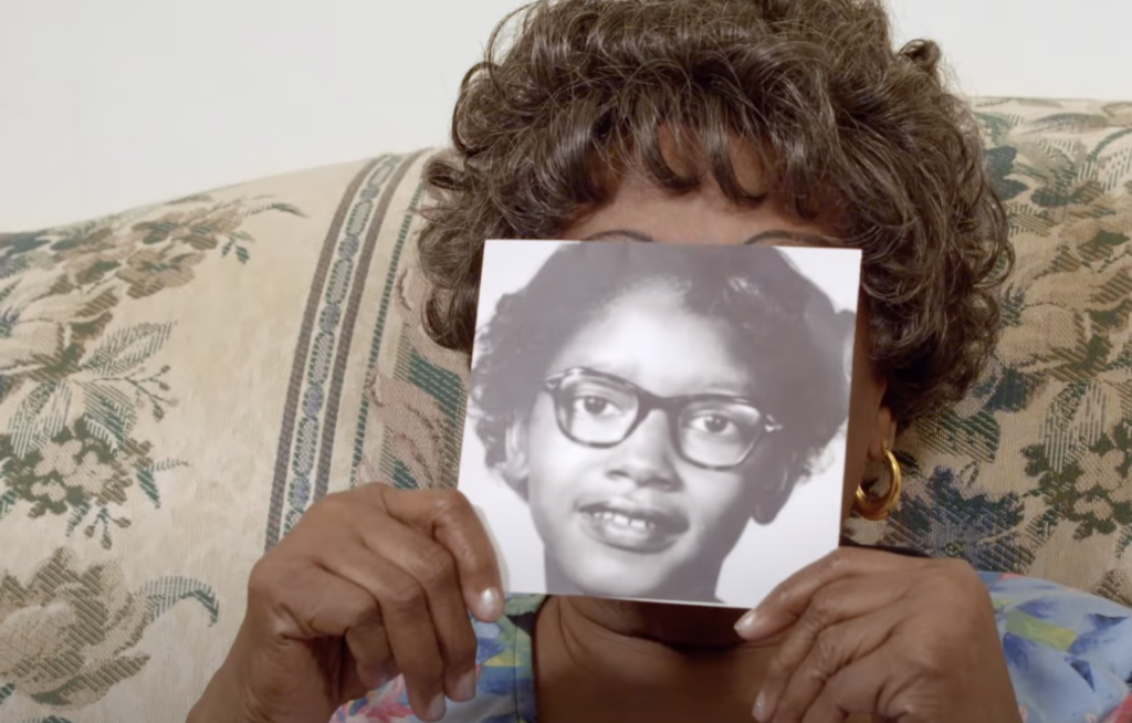 A person sitting on a floral-patterned couch holds up a black-and-white photo covering their face. The photo shows a young girl with glasses and curled hair. The person's hair and earrings are visible around the photo.