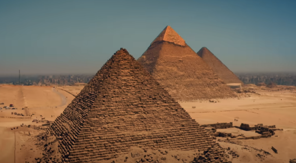 A sweeping view of the Giza pyramids under a clear blue sky. Three pyramids stand in the desert, the largest in the foreground. The city skyline is faintly visible in the background.