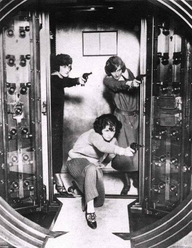 Three women with bobbed hairstyles and 1920s attire hold guns while standing in a large, open bank vault door. Each woman strikes a dynamic pose, one crouching in front, two standing behind her. The scene is black and white.