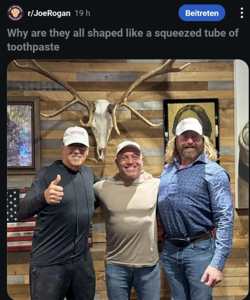 Three men wearing baseball caps stand smiling in front of a rustic wooden wall with a deer skull and antlers mounted on it. One man gives a thumbs-up, and there's an American flag on the side.