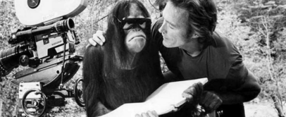 A man and an orangutan are on a film set. The orangutan is wearing sunglasses and holding a book. A film camera is nearby, and the man is leaning toward the orangutan, smiling. The background features trees. The image is in black and white.