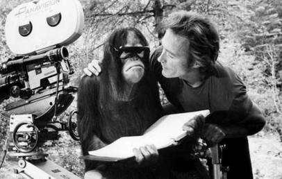 A man and an orangutan are on a film set. The orangutan is wearing sunglasses and holding a book. A film camera is nearby, and the man is leaning toward the orangutan, smiling. The background features trees. The image is in black and white.