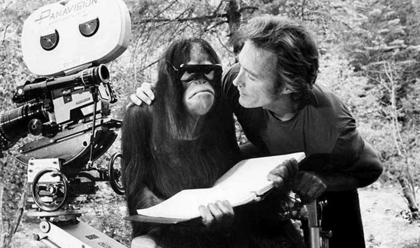 A man and an orangutan are on a film set. The orangutan is wearing sunglasses and holding a book. A film camera is nearby, and the man is leaning toward the orangutan, smiling. The background features trees. The image is in black and white.