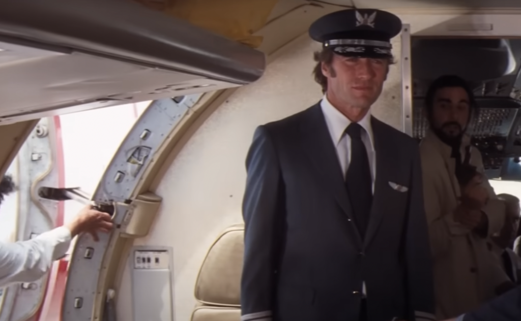 A pilot in uniform stands inside an airplane near the cockpit entrance. Another person in civilian clothing is visible in the background near the cockpit. A third person holds the door open from outside the plane.