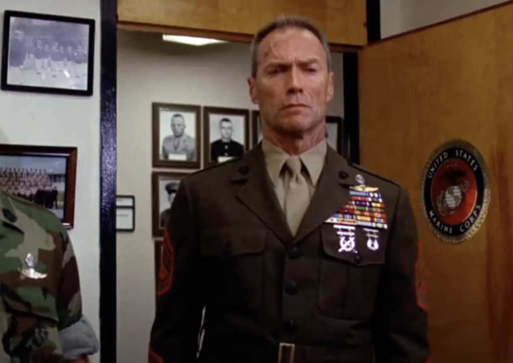 A stern-looking man in a military uniform stands indoors, adorned with numerous medals on his chest. Behind him are several framed black-and-white photos on the wall. A United States Marine Corps emblem is partially visible to his right.