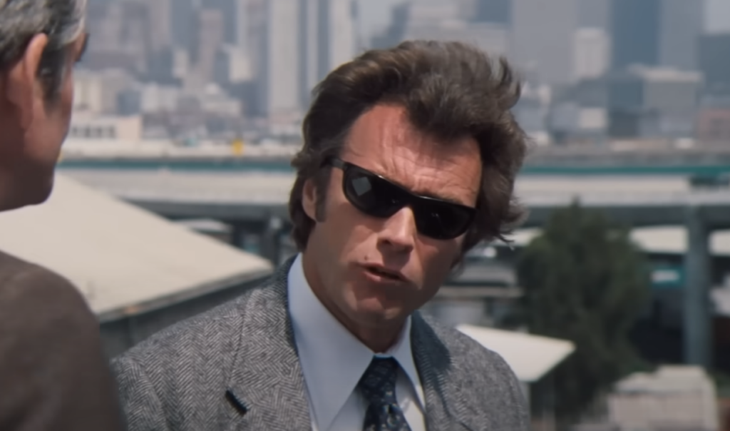 A man in a gray suit and dark sunglasses is talking to another person outdoors. The background shows a cityscape with buildings and highways.