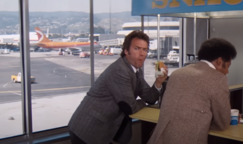 Two men in suits stand at an airport snack bar. One man is eating a sandwich and looking back, while planes and vehicles are visible through large windows in the background.