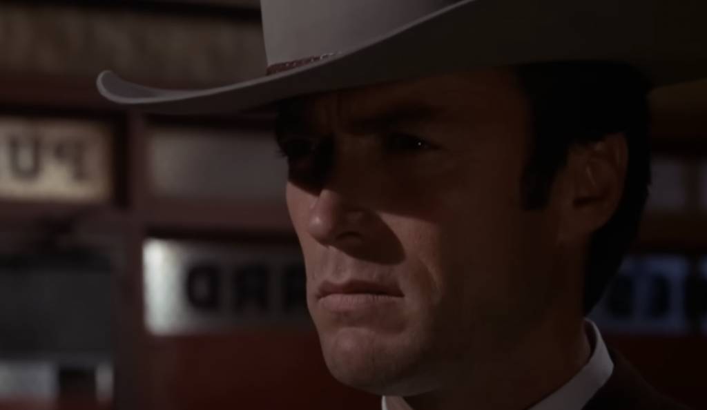 A person wearing a cowboy hat looks intently forward with a serious expression. The background is blurred, featuring indistinct signage or text. The lighting casts dramatic shadows on the person's face.