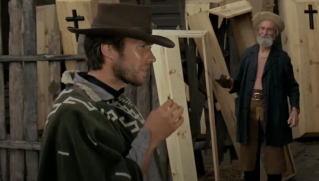 A man in a cowboy hat and poncho stands in the foreground, while an older man in a long coat holds a flask in front of coffins leaning against a wooden fence in the background.