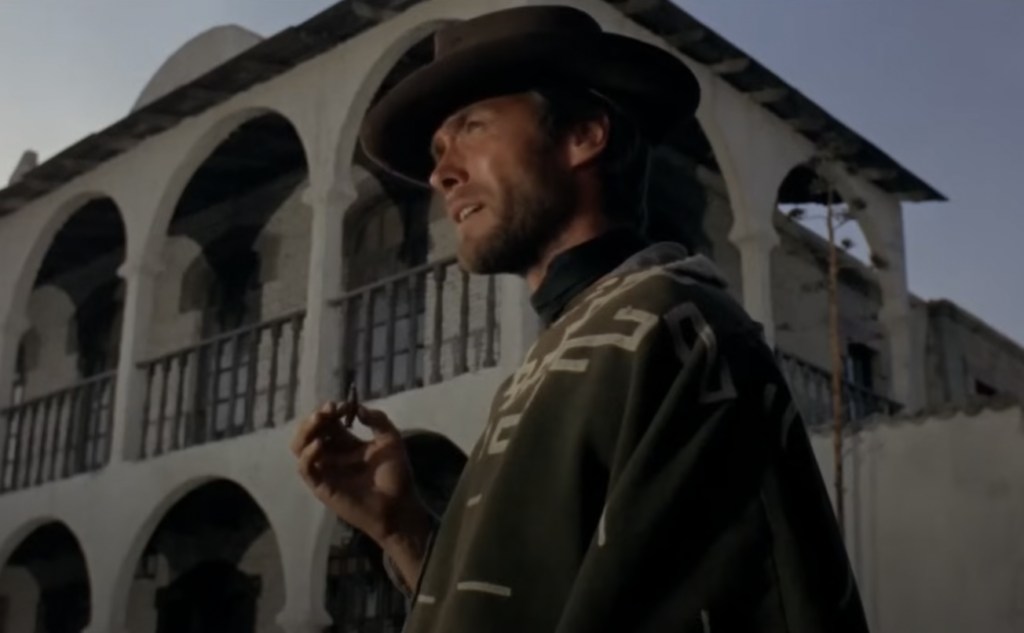 A person in a cowboy hat and poncho stands in front of a rustic building with archways, holding a small object. The sky is clear, and the building has balconies on each level. The scene has a Western-style ambiance.