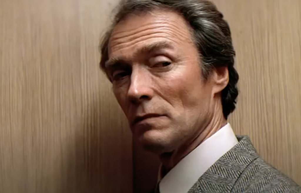 A man with gray hair and a serious expression stands in front of a wooden background, wearing a gray suit jacket and white shirt. He is looking over his shoulder towards the viewer.