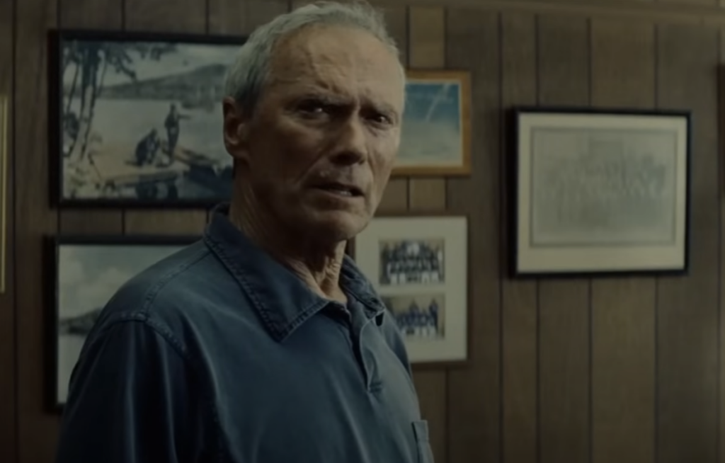 Elderly man with short gray hair wearing a dark shirt stands indoors in front of a wooden paneled wall adorned with several framed photographs. He looks slightly to the side with a serious expression.