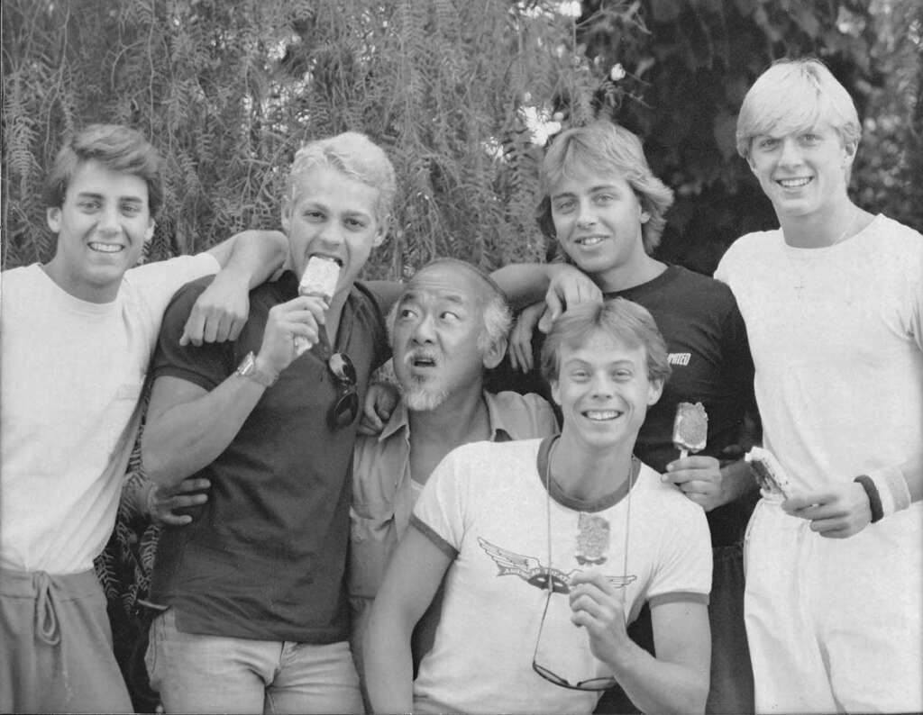 A group of six people, five young men and one older man, stand together outdoors. The young men hold ice cream bars and smile at the camera, while the older man looks up with a playful expression. They appear relaxed and happy.