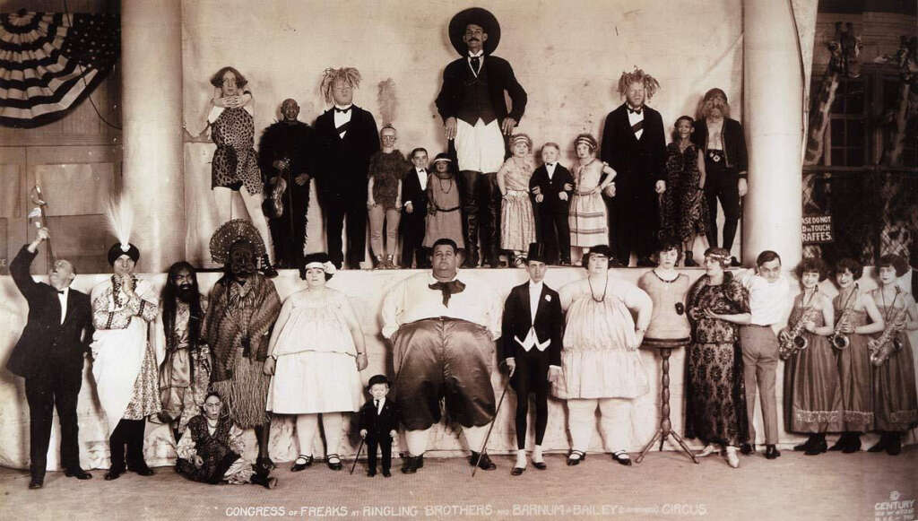 A vintage black-and-white photo of a circus sideshow group. Various performers, including those with unique physical traits, are posed in rows. The backdrop features a marquee with circus banners and flags.