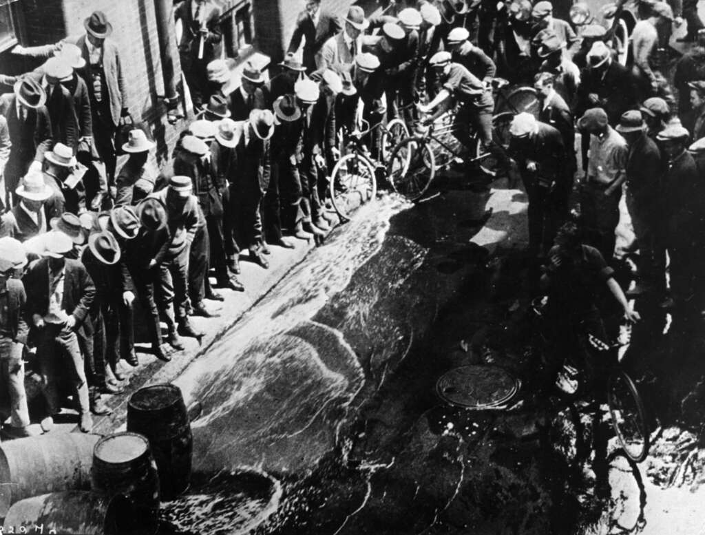 A group of men in hats stand along the sides of a street watching a stream of liquid flow from a large barrel. Some hold bicycles. The scene appears to be from an early 20th-century urban setting.