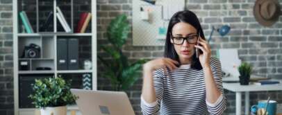 A woman wearing glasses and a striped shirt is talking on the phone while gesturing with her hand. She is sitting at a desk with an open notebook and a laptop. Behind her are shelves, a plant, and a brick wall with decor.