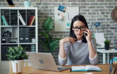 A woman wearing glasses and a striped shirt is talking on the phone while gesturing with her hand. She is sitting at a desk with an open notebook and a laptop. Behind her are shelves, a plant, and a brick wall with decor.