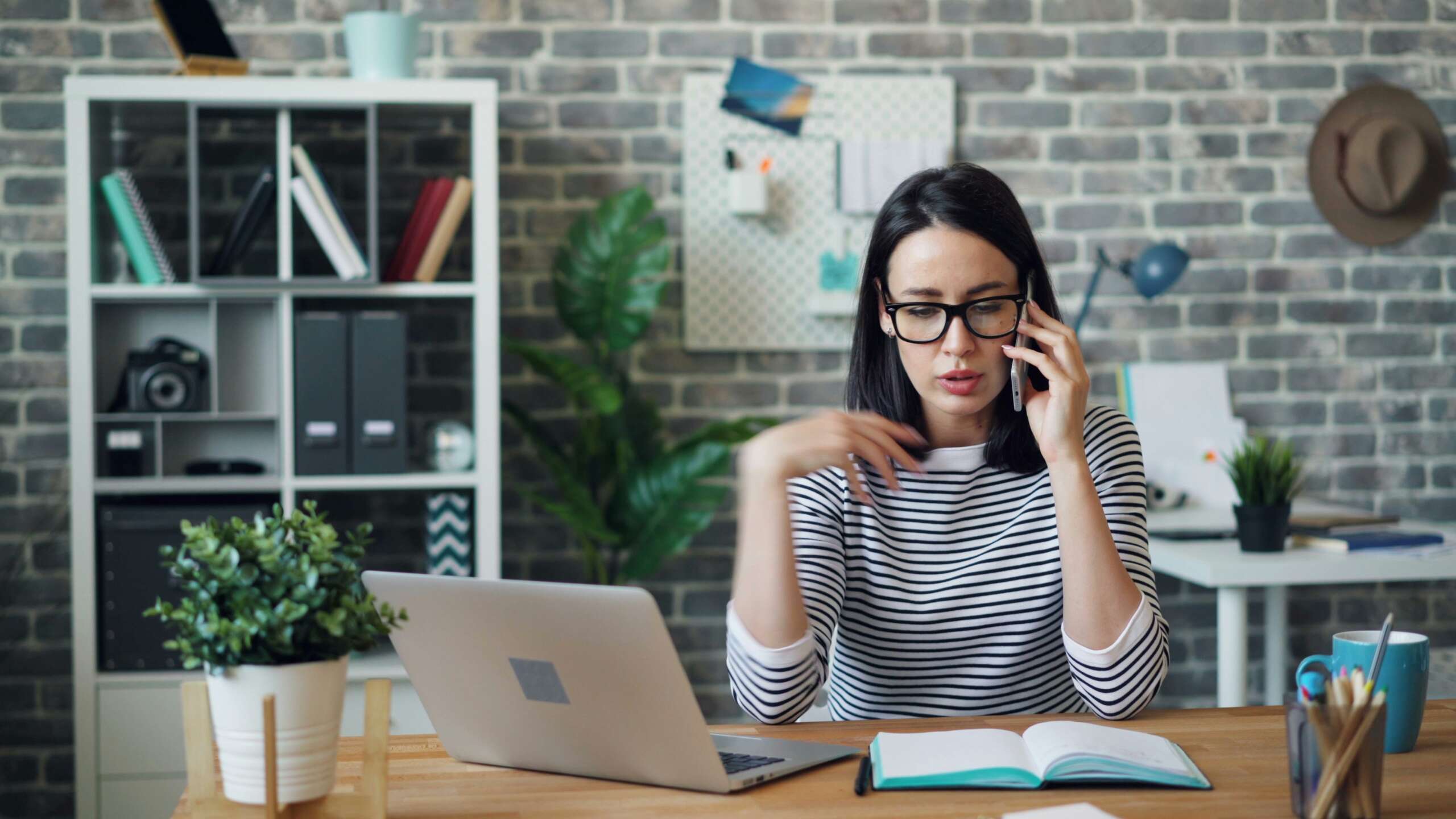 A woman wearing glasses and a striped shirt is talking on the phone while gesturing with her hand. She is sitting at a desk with an open notebook and a laptop. Behind her are shelves, a plant, and a brick wall with decor.