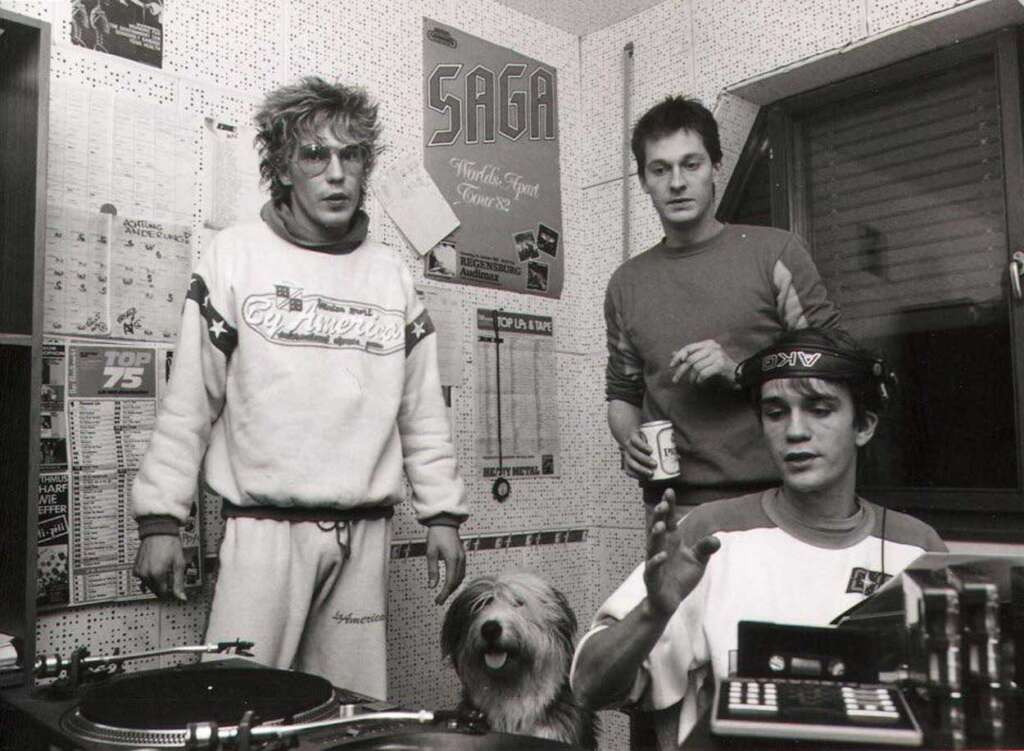 Three men in a small room with music equipment, a dog at their feet. Posters and charts cover the walls. One man in a tracksuit stands beside the DJ setup, another holds a can, and the third is seated, adjusting controls.