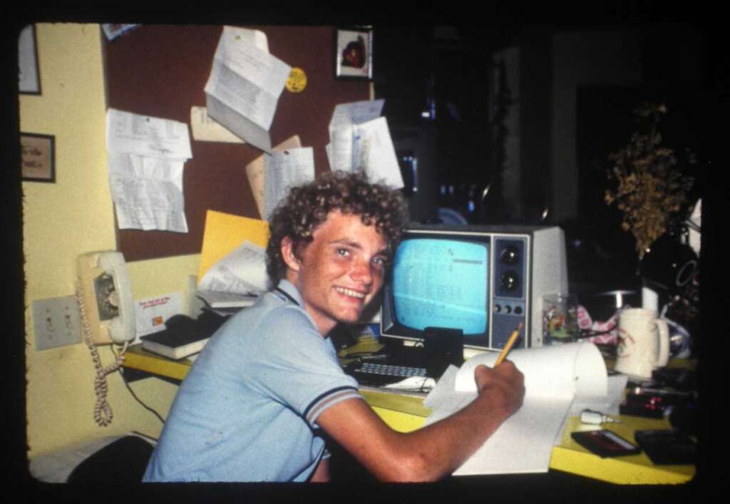 A person with curly hair smiles while sitting at a desk, writing on paper. A vintage computer with text on the screen is in front. The desk is cluttered with papers, and a bulletin board with notes is in the background.