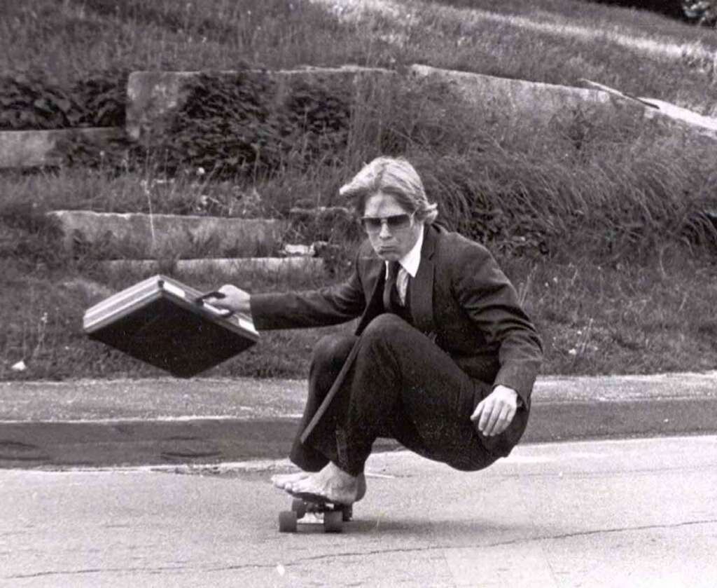 A person in a suit and sunglasses is crouching on a skateboard, holding a briefcase, while skateboarding on a street. The background shows grass and stone steps.