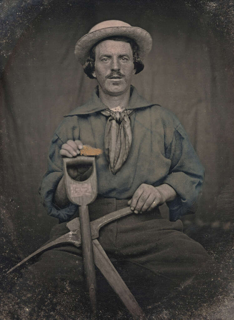 A vintage photograph of a man wearing a hat and holding a saw and pickaxe. He has a mustache and is dressed in a long-sleeve shirt with a scarf tied around his neck. The image has a sepia tone, giving it an antique appearance.