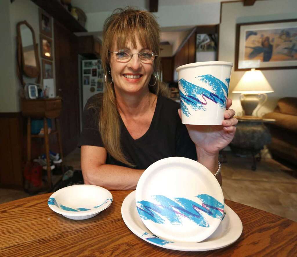 A woman smiling and holding a white cup with a blue and purple wave design. In front of her on a wooden table are a bowl and two plates with the same design. The background shows a cozy living room.