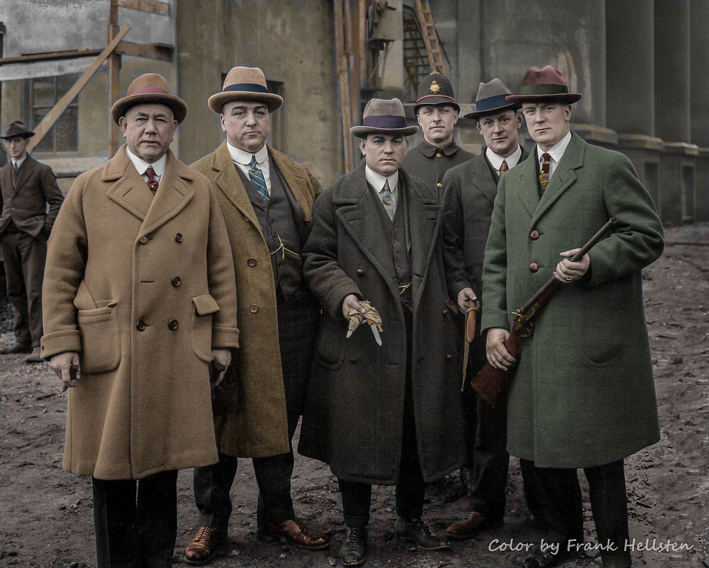 A historical photograph of six men in long coats and hats standing together. The group includes two men holding guns, and one in a police uniform. The background shows a building under construction. The image is colorized.